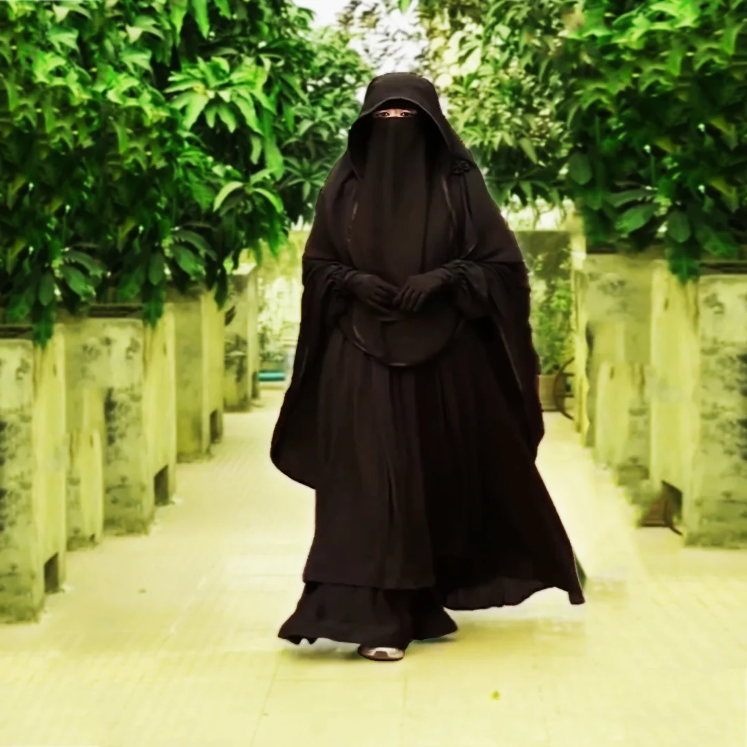 A young girl wearing a black niqab dress walks towards the cameraman. Her face is covered with a black cloth.