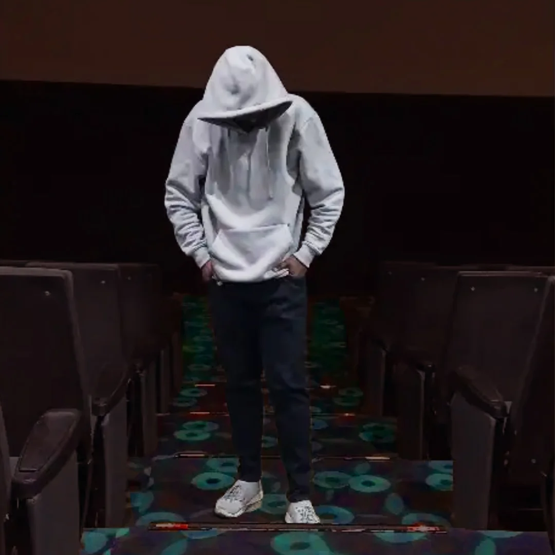 A unconfident young boy wearing white hoodie in a hall. His hands are inside his jeans and looking downwards as if he is sad.