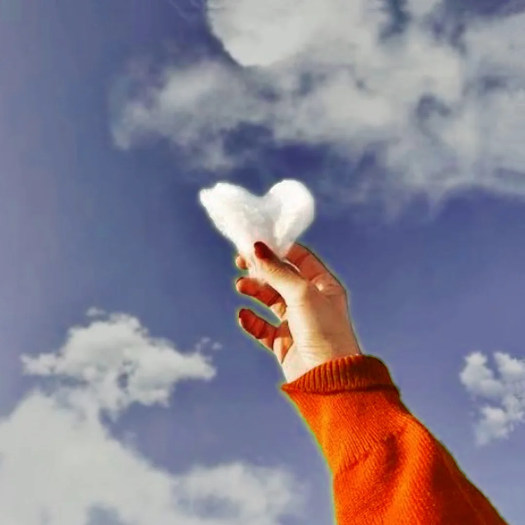 An image of a hand and a sky filled with clouds. A hand is holding clouds in the shape of love.