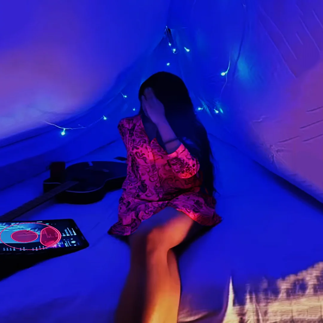 A gorgeous teen girl sitting on the bed at a party. She is wearing a short coat while her face is covered with her hand. The room is light up in blue and pink. At the same time, Guiter and iPad Pro on her side