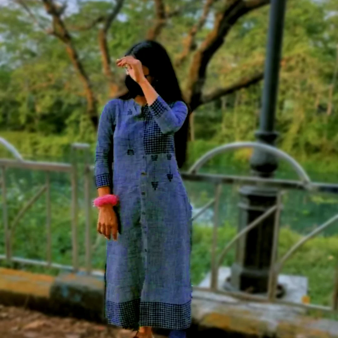 A young teen shy girl posing in a park surrounded by trees, water and greenery. She is wearing a blue salwar while her face is covered with a mask and hand.